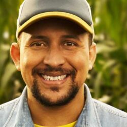 homem de meia-idade sorrindo. Ele está com um boné cinza de aba amarela e uma camisa jeans. Ao fundo, desfocada, uma plantação